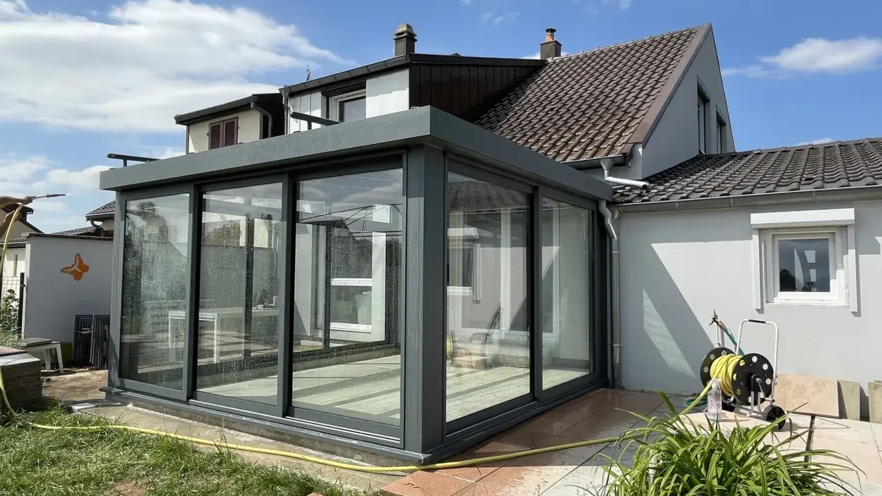 Une véranda moderne en verre et métal, prête pour l'assurance. Le soleil brille sur le jardin et la maison.