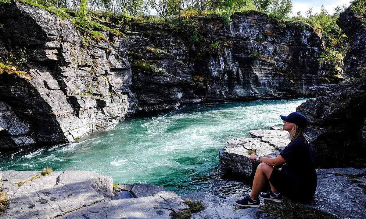 En kvinna sitter vid en forsande turkos flod i en klippig ravin. Vacker natur, kanske en vy över var ligger Abisko.