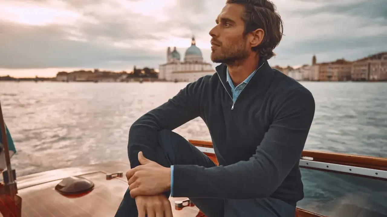 A man in mens dressy casual attire, a navy quarter-zip sweater over a blue collared shirt, sits on a boat with Venice in the background.