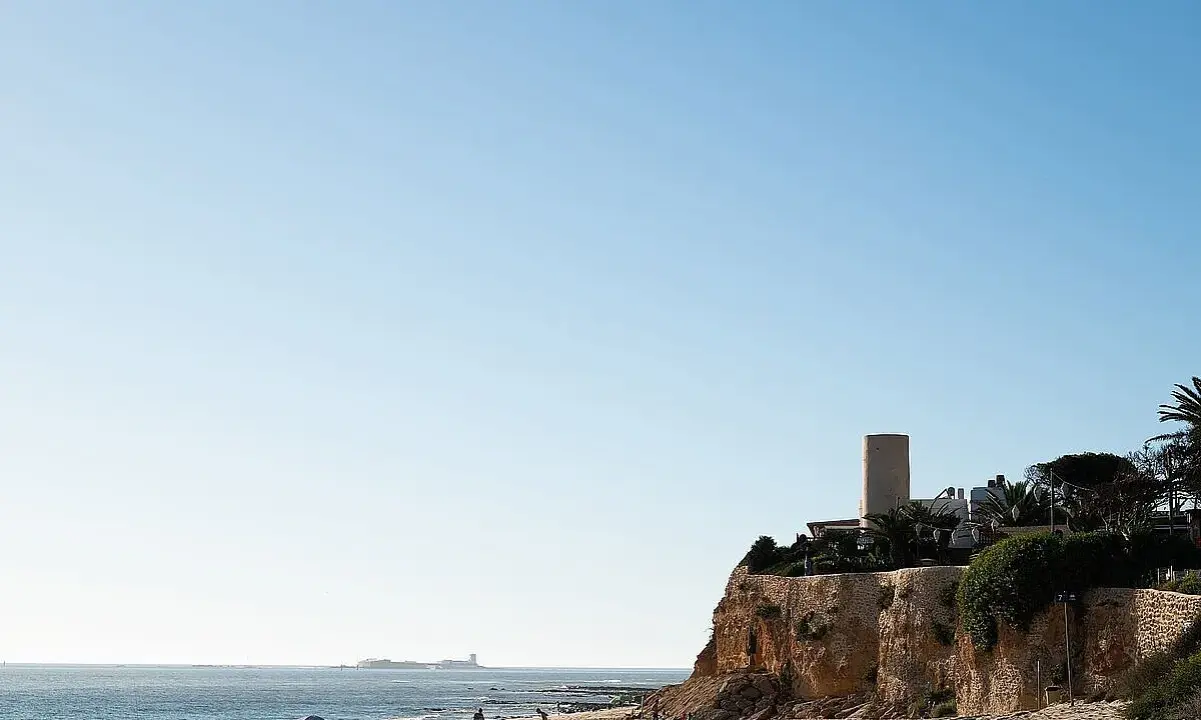 Sonniger Tag am **Playa La Barrosa**. Menschen genießen den Strand, während im Hintergrund eine Festung am Meer thront.