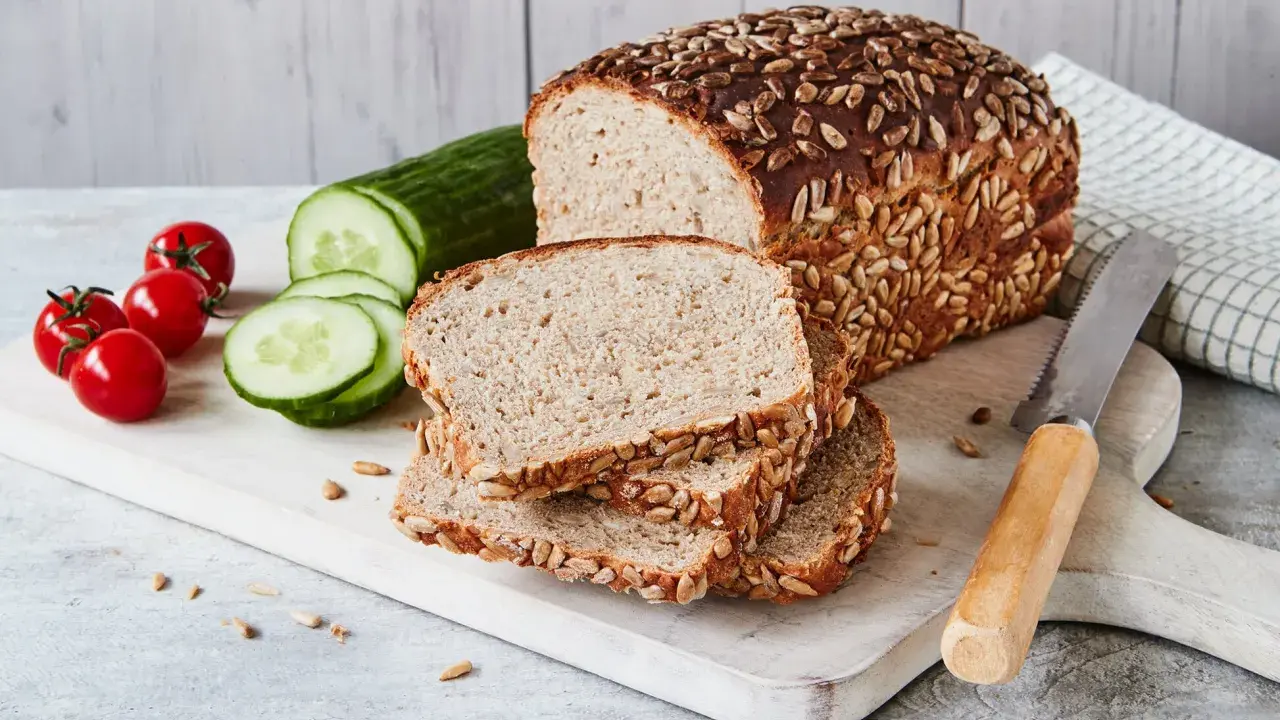 Frisch gebackenes Sonnenblumenbrot, zubereitet im Thermomix, liegt auf einem Holzbrett neben Tomaten und Gurkenscheiben.