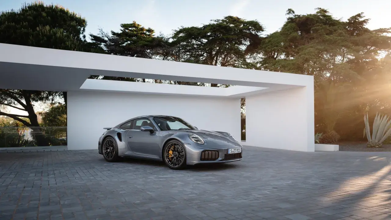 Silberner Porsche Turbo S, bereit für den 0-100 Sprint, parkt vor moderner Architektur im Sonnenuntergang.