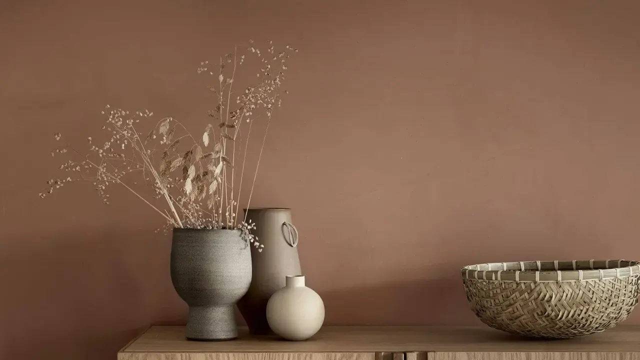 Tre vaser med torkade blommor och en flätad korg står på en träbyrå. En av vaserna är grå och har en rustik textur, en annan är beige med ett handtag och den minsta är rund och ljus.