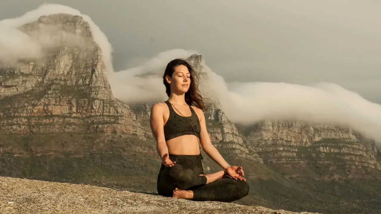 Kvinna i lotusställning på en klippa, med berg och moln i bakgrunden. Hon utövar buddhistisk meditation för inre lugn.