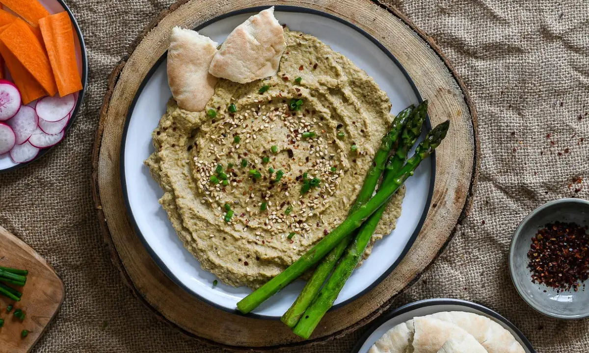 Hummus de berenjena cremoso, decorado con sésamo y espárragos, acompañado de pan de pita y crudités.