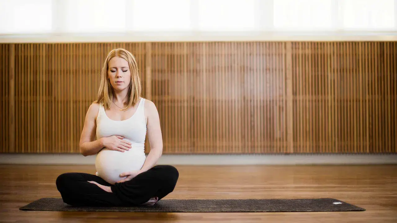 Gravid kvinna med slutna ögon sitter i lotusställning på en yogamatta. Hon håller sin mage, en lugn stund av gravid yoga.