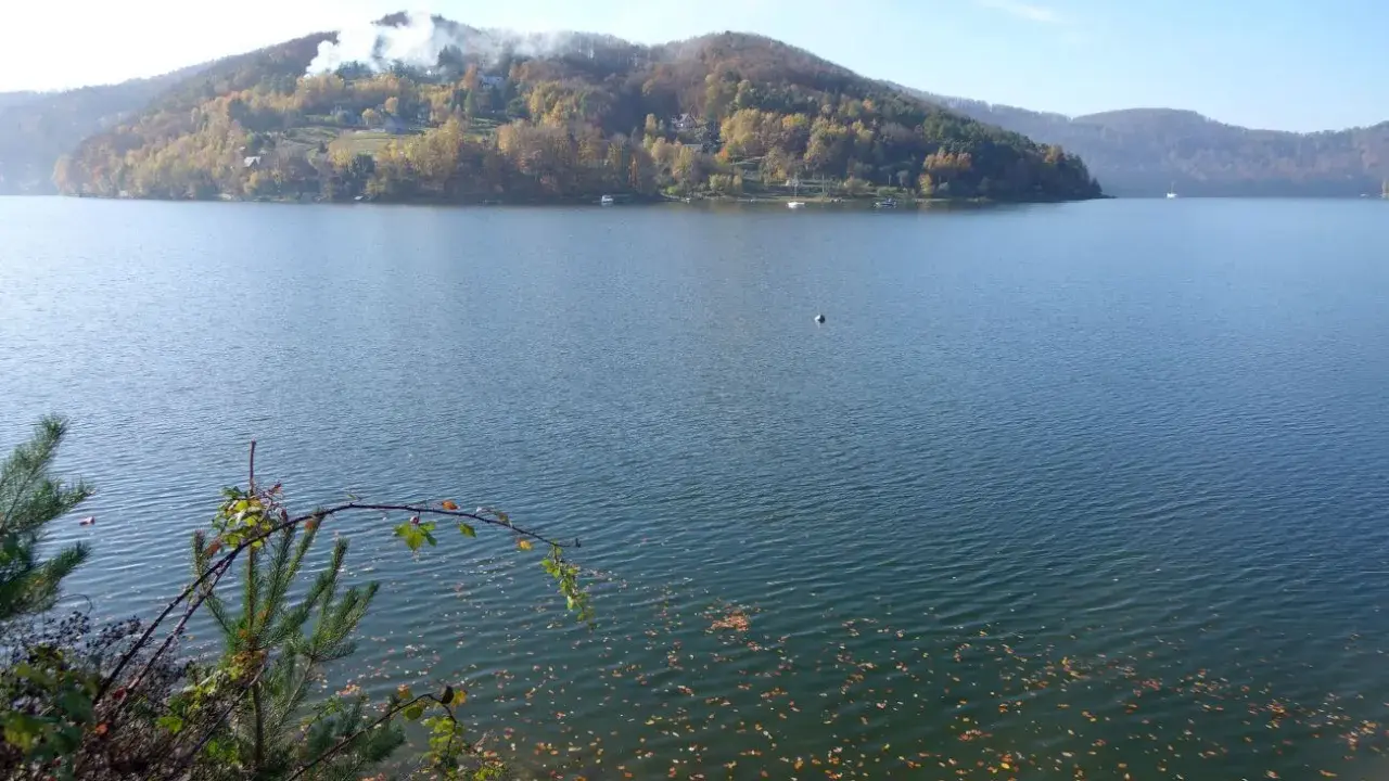 Jesień nad zalewem rożnowskim. Woda spokojna, na brzegu opadłe liście. W oddali wzgórza porośnięte jesiennymi barwami.