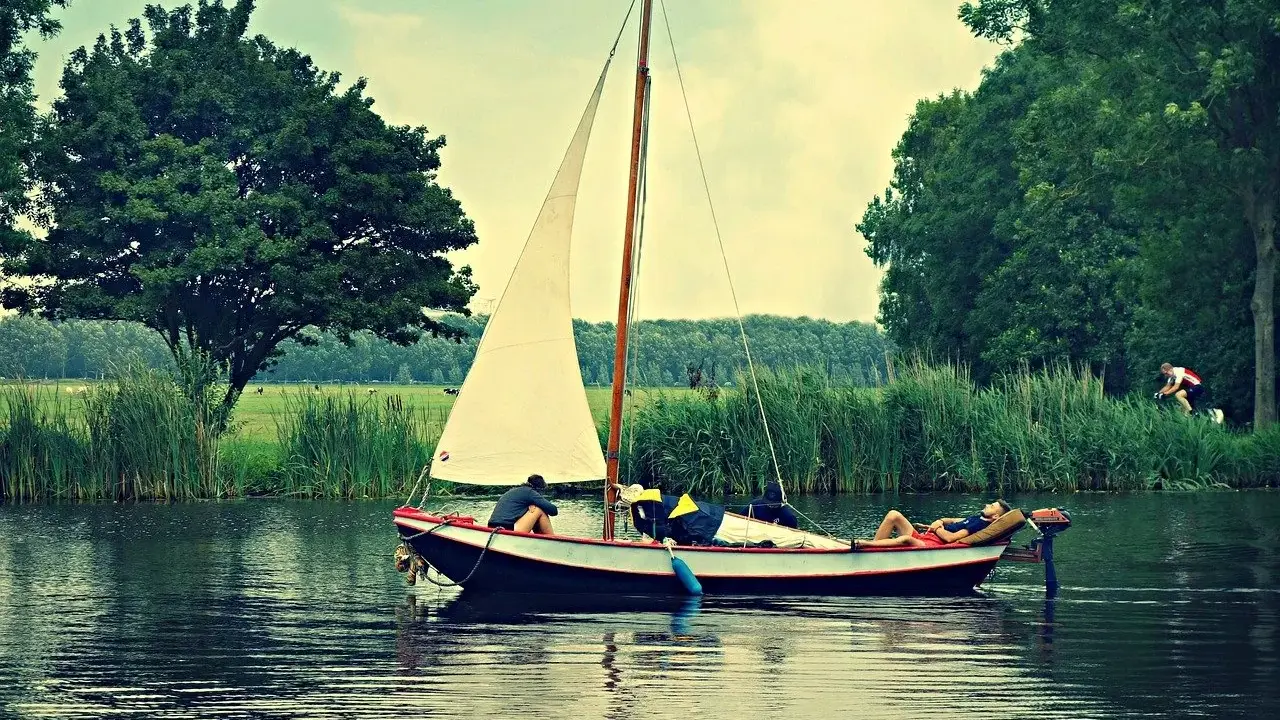 Un petit type de bateau à voile navigue sur un canal bordé d'arbres. Une personne se repose sur le pont, une autre est assise.