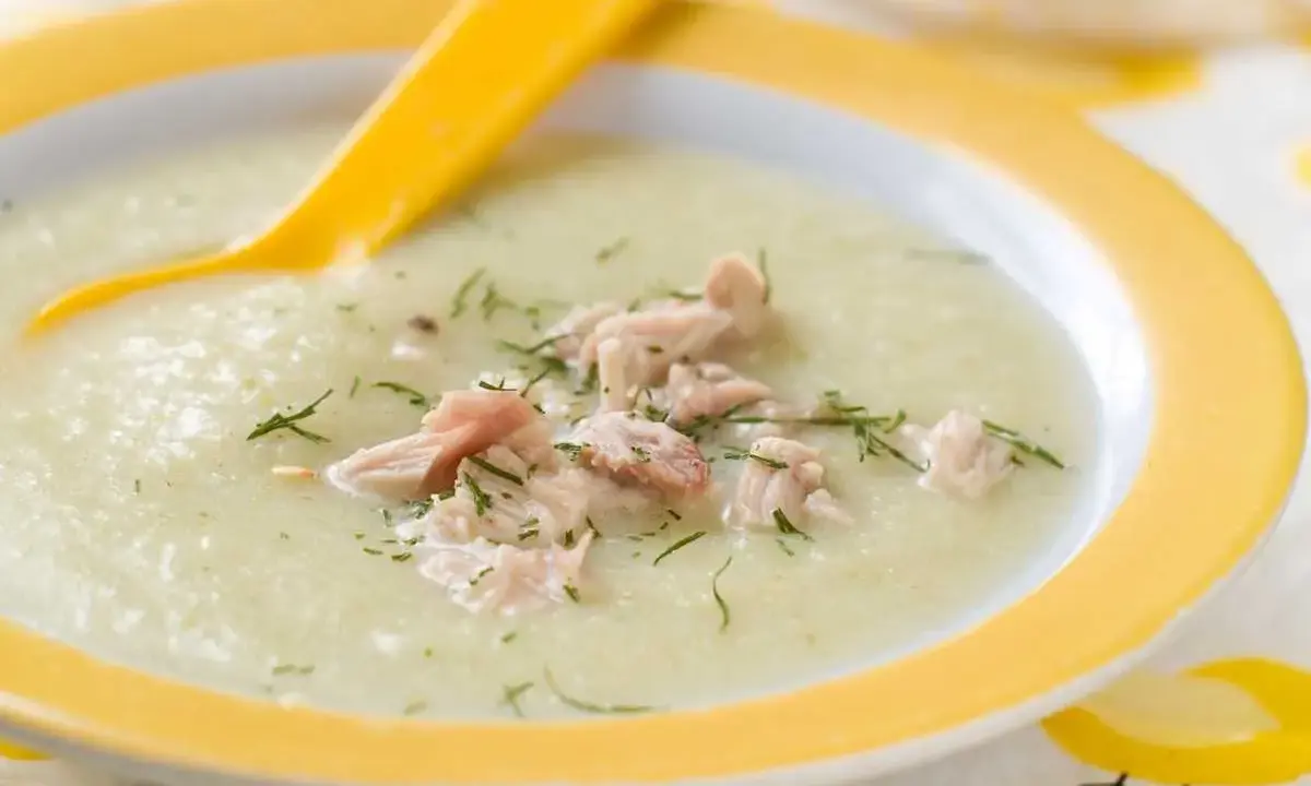 Un cremoso puré de verduras con pollo desmenuzado y eneldo, servido en un plato amarillo con una cuchara.