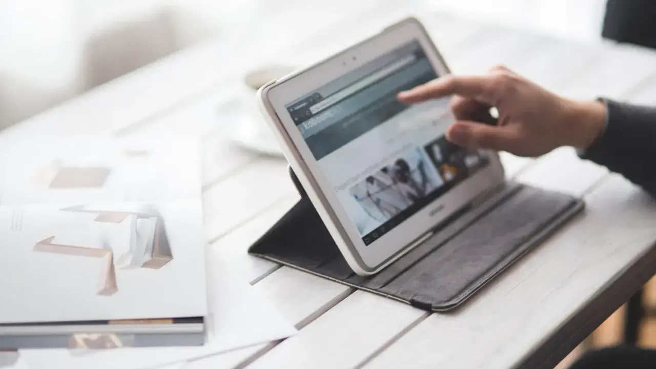 Dłoń wskazuje na ekran tabletu, który wyświetla stronę internetową. Obok leży otwarta książka z grafikami. W tle widać filiżankę. To idealne miejsce do pracy nad projektem, gdzie każdy element, nawet **padding CSS**, ma znaczenie.