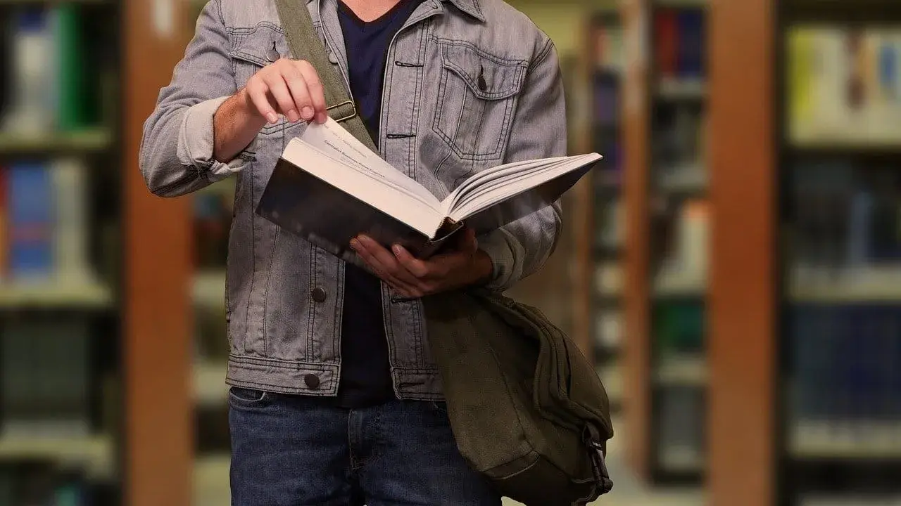 Student w dżinsowej kurtce przegląda książkę w bibliotece. W tle widać regały z książkami.
