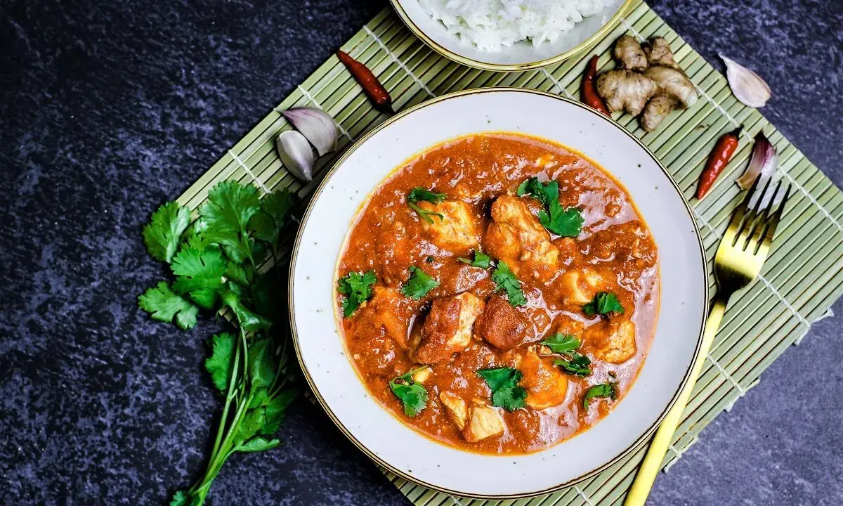 Ein Teller mit Hähnchen-Curry in cremiger **masala sauce**, garniert mit Koriander. Daneben Reis, Ingwer, Knoblauch und Chilis.