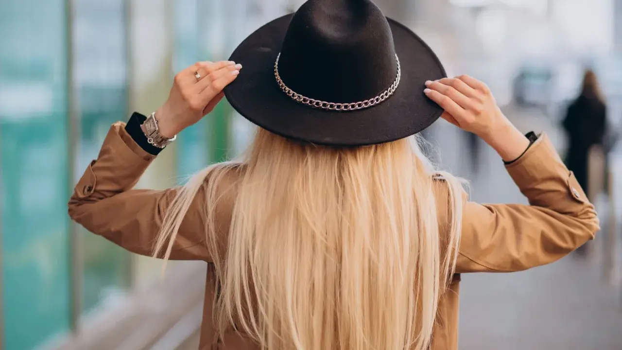 Blondynka w beżowym płaszczu i czarnym kapeluszu z ozdobnym łańcuszkiem. Pokazuje, jak nosić kapelusz, poprawiając go na głowie.