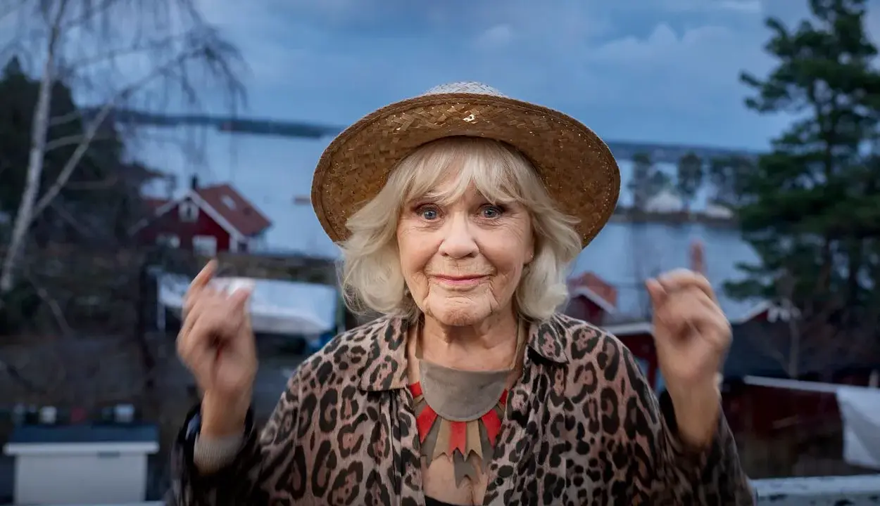 Christina Schollin, med sin karaktäristiska hatt och leopardmönstrade jacka, ser ut att berätta en historia från sina filmer och tv-program.
