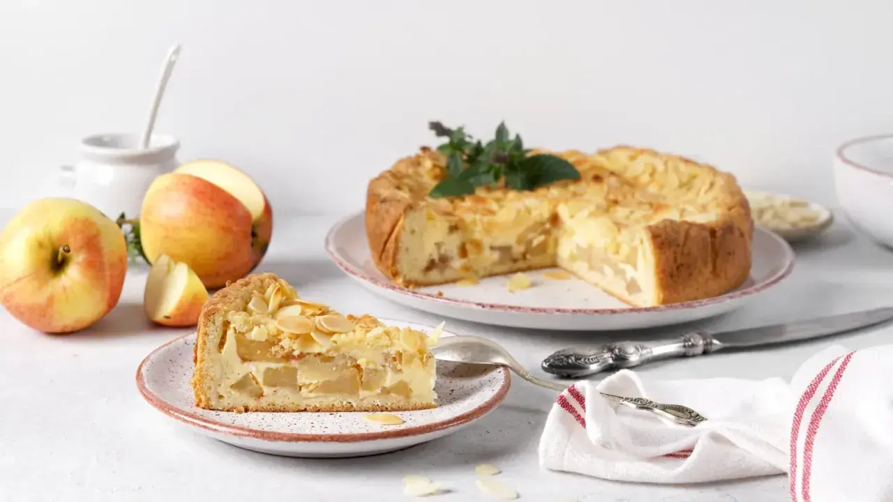 Un délicieux gâteau aux pommes, garni d'amandes effilées, est présenté avec une part déjà coupée. Des pommes fraîches sont disposées à côté.