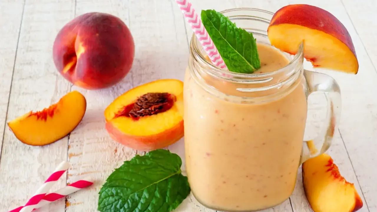 Erfrischender Pfirsich Smoothie im Glas, garniert mit Minze und Pfirsichspalten. Dazu frische Pfirsiche und Trinkhalme.