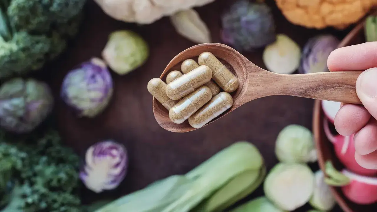 Łyżka z kapsułkami – witaminy na energię. W tle świeże warzywa: brokuły, brukselka, kapusta pak choi.