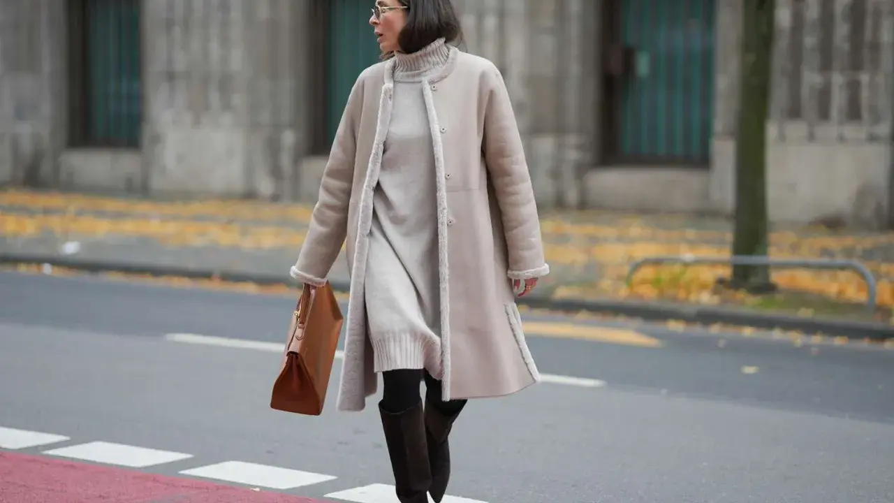 A woman demonstrates how to layer a dress for winter with a cozy turtleneck dress, a shearling coat, and stylish boots.