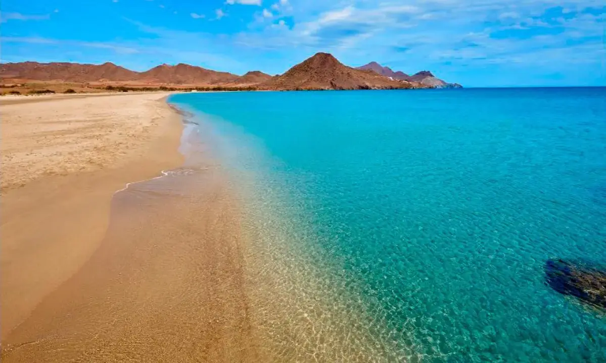 La **playa de los Genoveses** se extiende con arena dorada y aguas cristalinas de color turquesa, bajo un cielo azul con nubes blancas y montañas áridas al fondo.