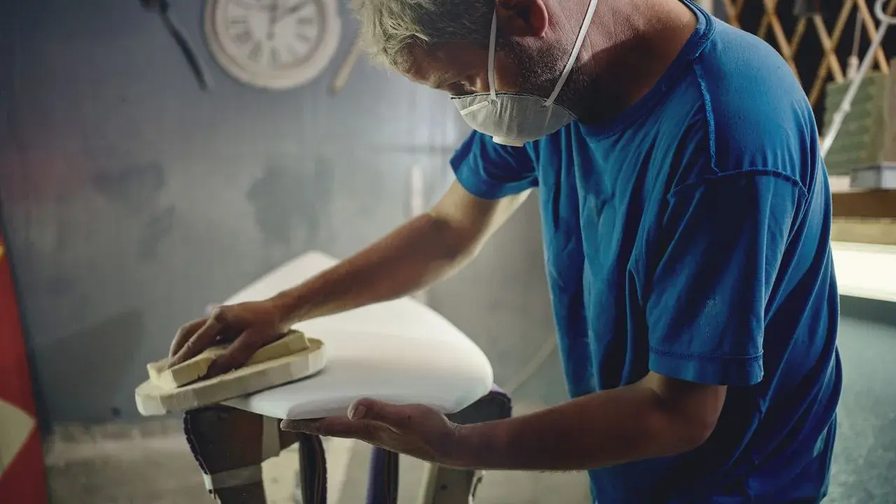 Artisan masqué ponçant une planche de surf blanche, étape cruciale avant de faire de l'époxy.