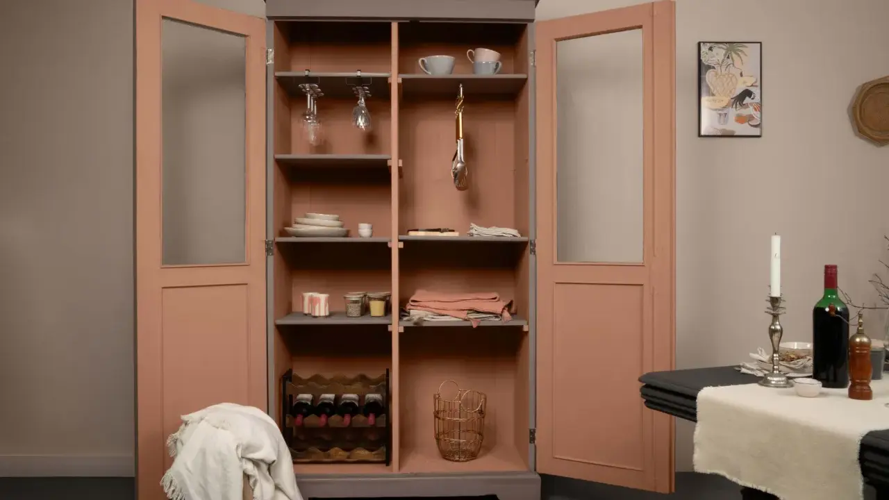 Armoire vitrée avec un relooking meuble 2 couleurs : gris anthracite et rose poudré. Elle contient vaisselle, verres, bouteilles de vin et objets déco.