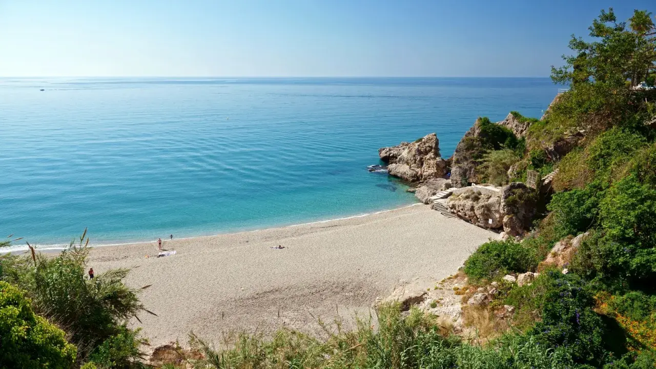 Playa de Burriana: Ein sonniger Strand mit türkisfarbenem Wasser, Felsen und üppiger Vegetation. Einige Menschen genießen die Ruhe.
