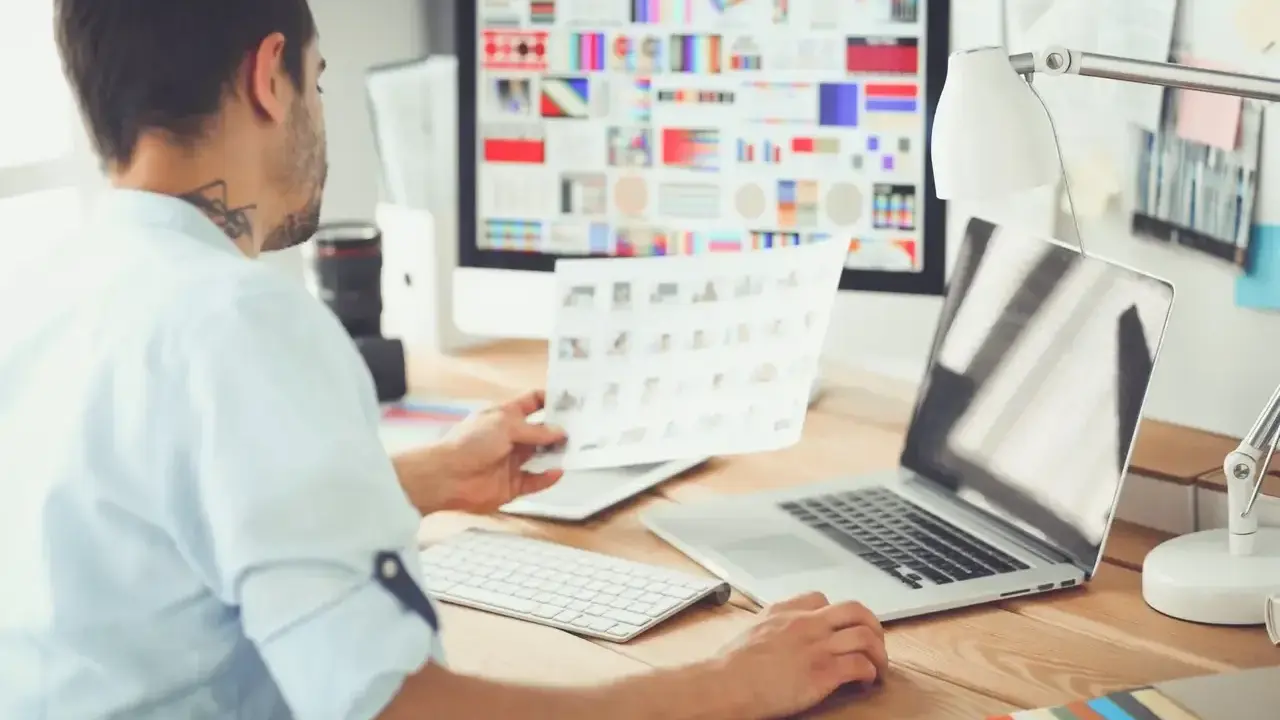 UX UI designer pracuje przy biurku, analizując wydruki i ekran komputera z paletą barw.