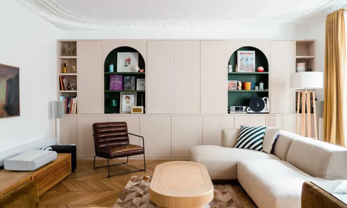 Un salon moderne avec un canapé beige, une chaise en cuir marron et des étagères murales roses. Quelle couleur pour une grande pièce à vivre ? Le vert émeraude des niches apporte une touche audacieuse.
