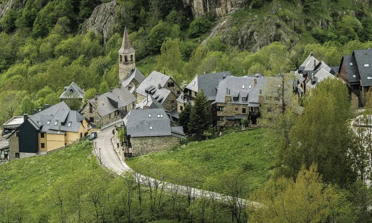 Pueblos del Valle de Arán, con casas de piedra y tejados grises, se asoman entre la vegetación verde y un camino serpenteante.