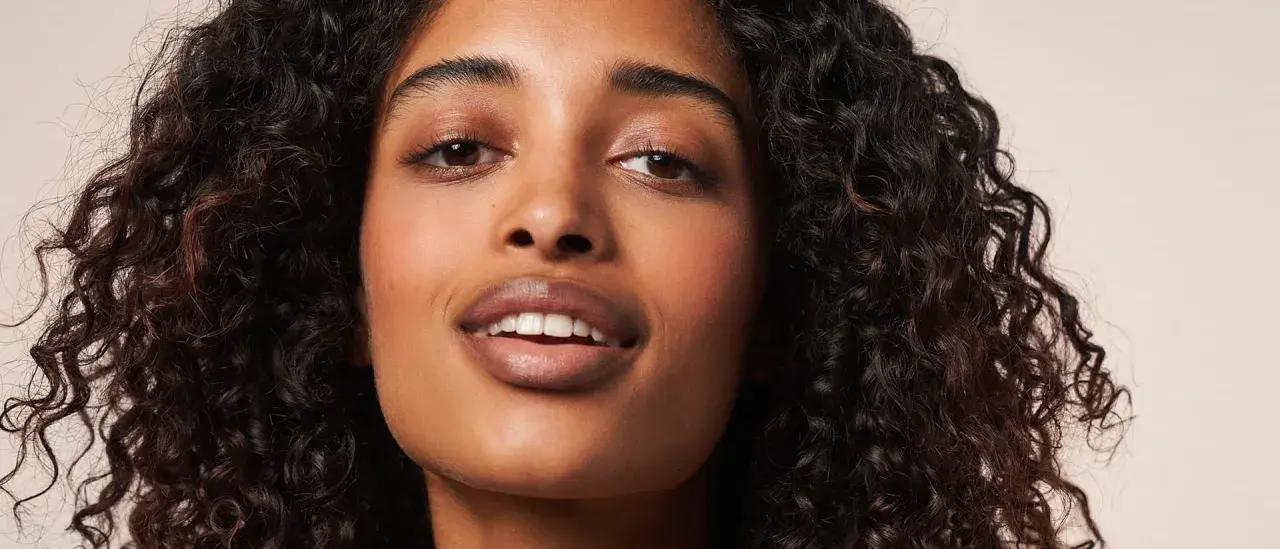 Ein Porträt einer Frau mit voluminösen, dunklen Locken, die die Curly Girl Methode perfektioniert. Ihre Haut strahlt, ihre Augen funkeln.