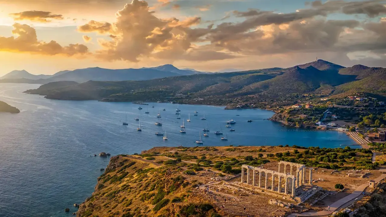 Rovine del Tempio di Poseidone a Capo Sounion, con barche a vela che navigano nel mare blu sotto un cielo dorato al tramonto.