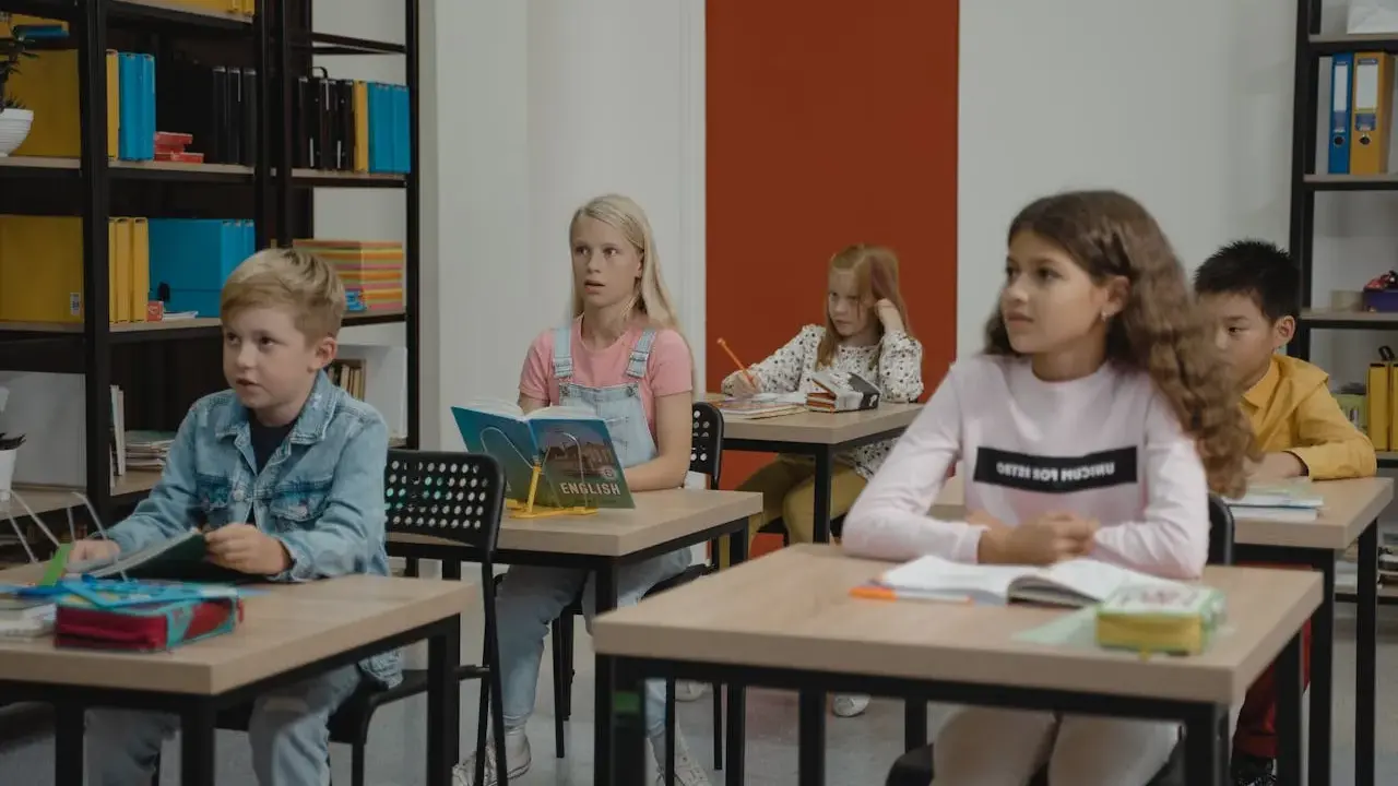 Dzieci siedzą w klasie, skupione na lekcji. Chłopiec w dżinsowej kurtce, dziewczynka z książką po angielsku, a z tyłu inne dzieci. Wszyscy mieli nadzieję, że lekcja szybko się skończy.