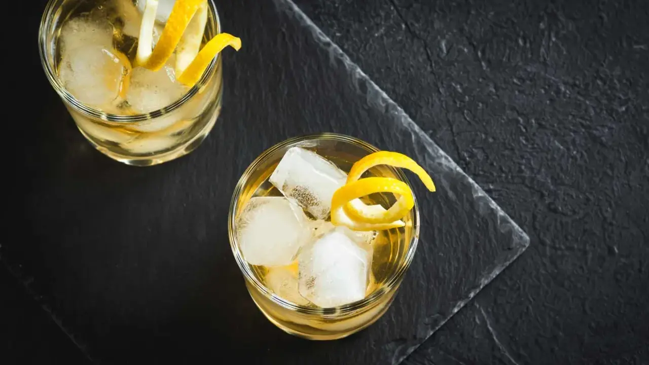 Zwei erfrischende Whiskey Cocktails mit Eis und Zitronenzesten auf dunklem Stein.