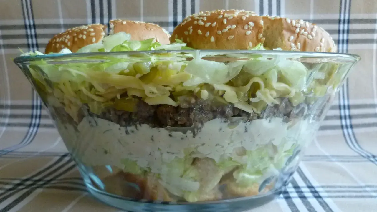 Warstwowa sałatka big mac w szklanej misie, z bułkami na wierzchu.