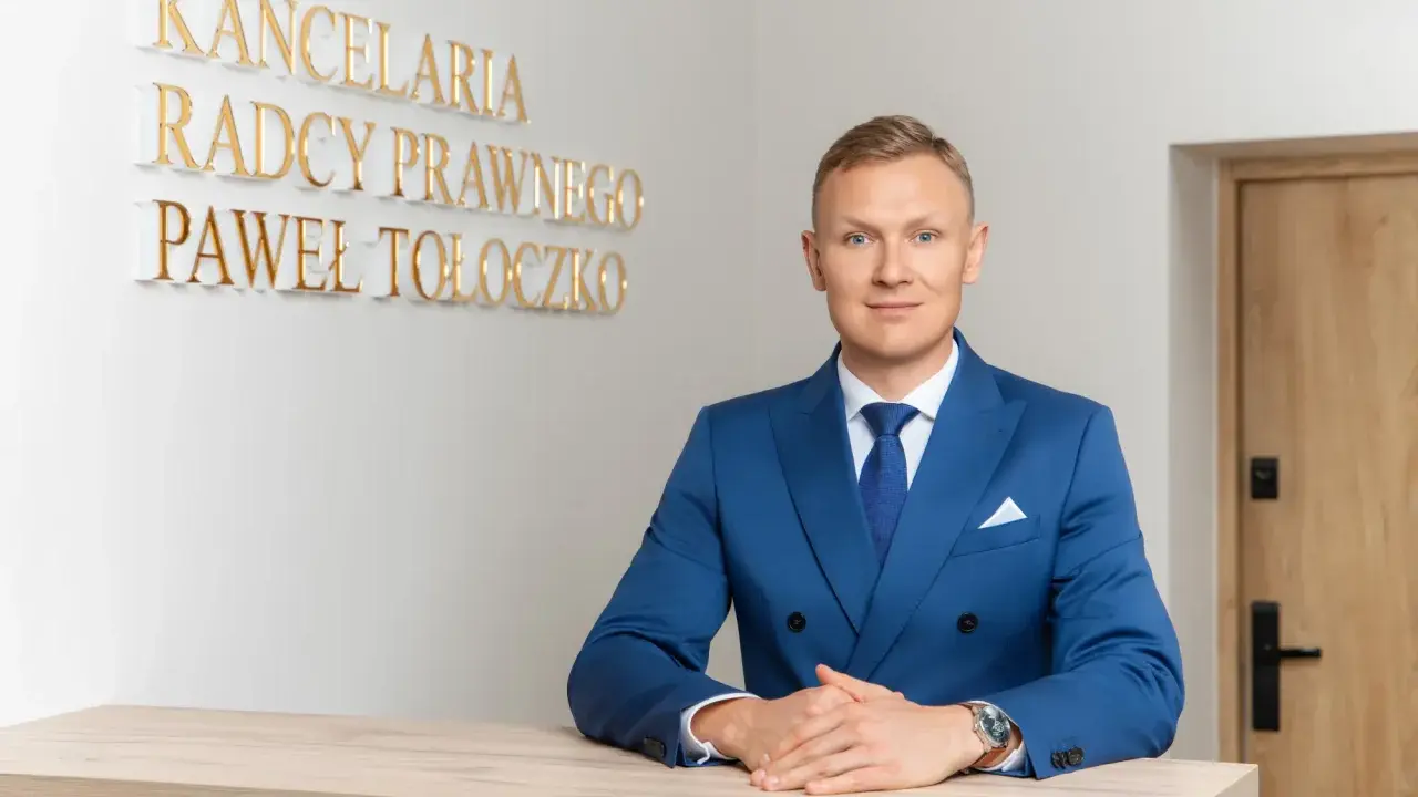 Mężczyzna w niebieskim garniturze przy biurku, za nim napis "KANCELaria Radcy Prawnego Paweł Tołoczko".