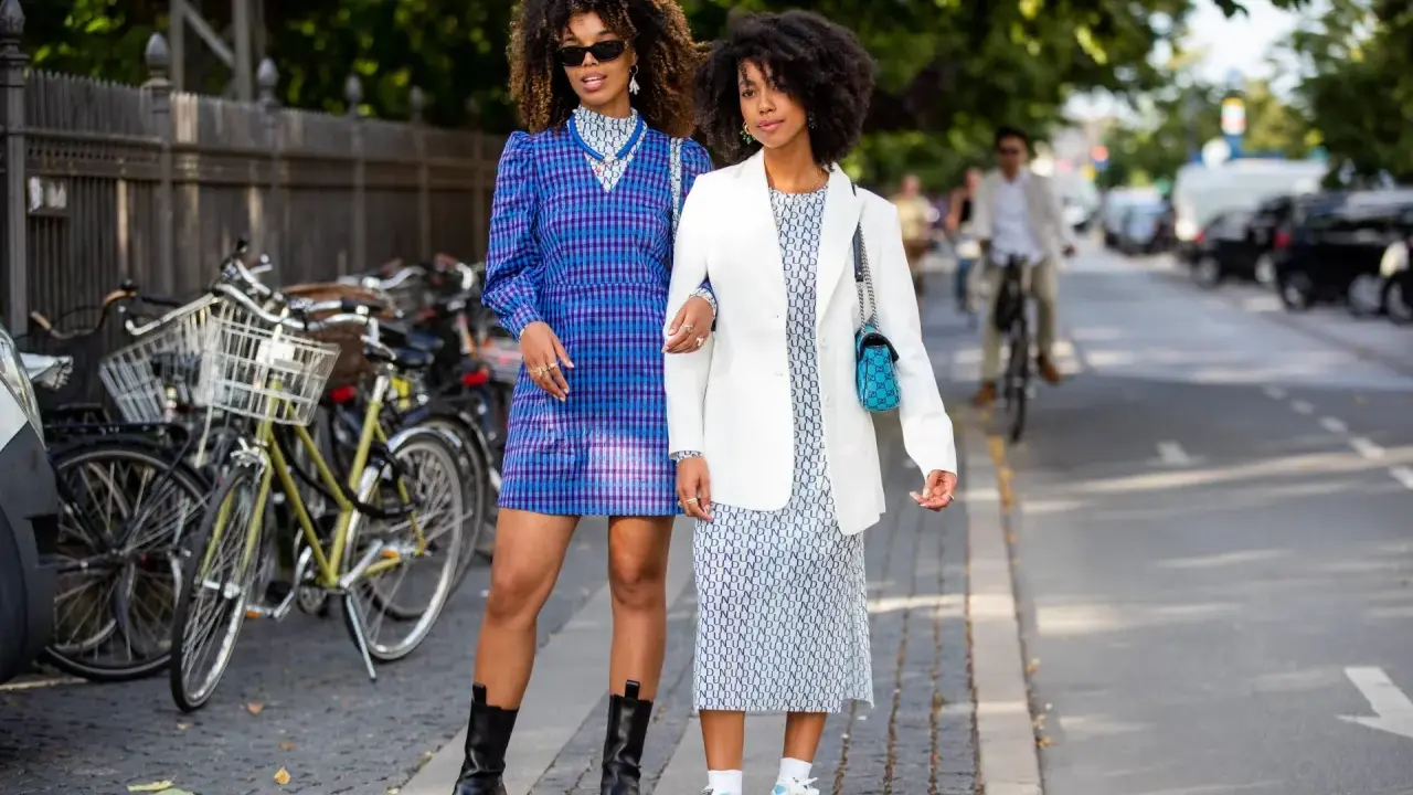Two women showcase their spring chic dress code. One wears a blue plaid dress and chunky boots, the other a white blazer over a patterned dress and sneakers.