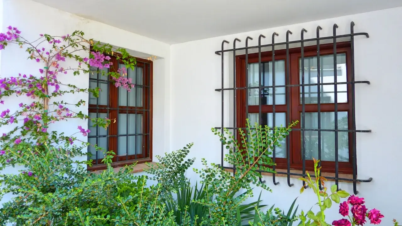 Dos ventanas con barrotillo de hierro y marcos de madera, rodeadas de vegetación y flores moradas.