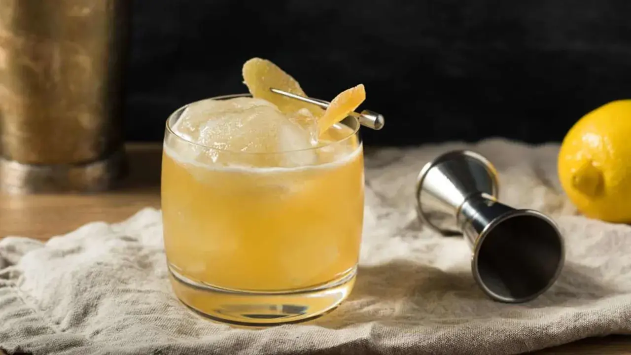 En gyllene whiskey drink med is och citronskal, redo att avnjutas. Bredvid står en jigger och en citron.