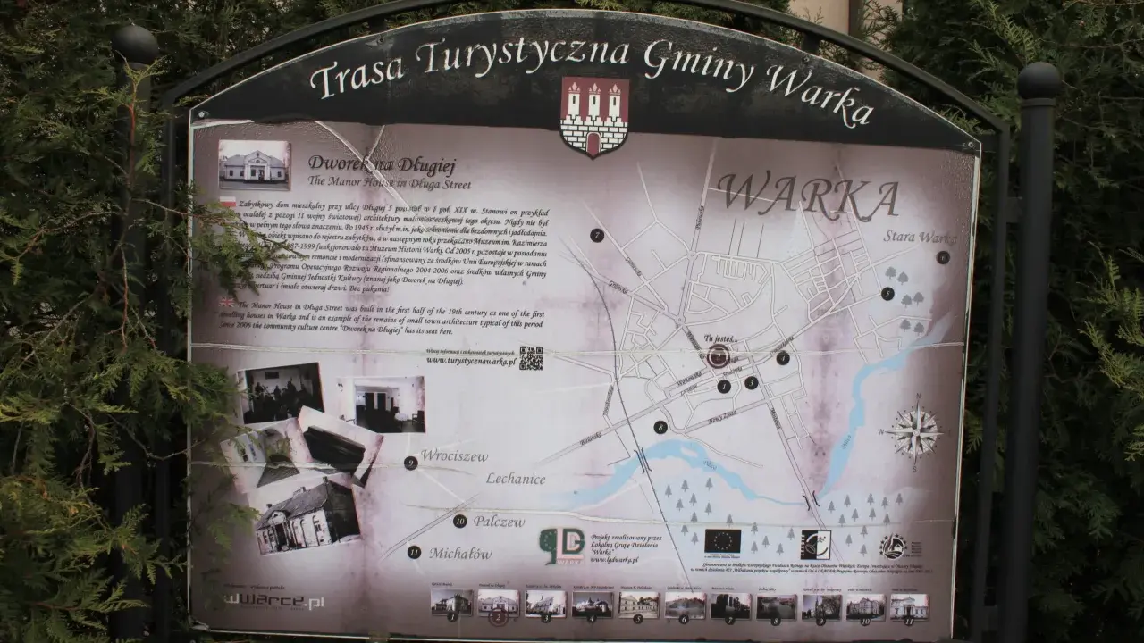 Mapa turystyczna gminy Warka z zaznaczonymi zabytkami, w tym Dworkiem na Długiej. W tle drzewa, a na dole strony logo "Warka" i adres strony.