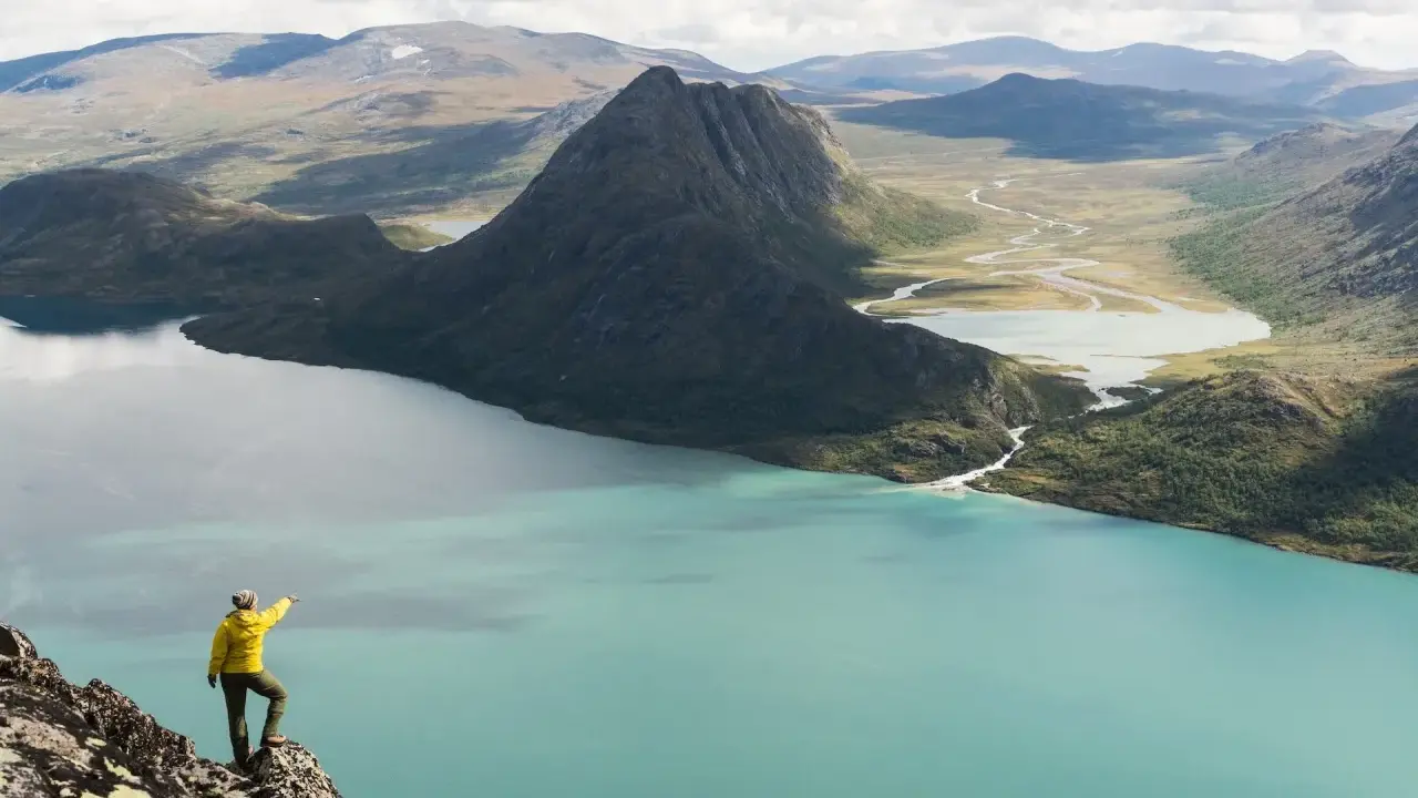 En person i gul jacka pekar ut över en turkos sjö och berglandskap under en Jotunheimen vandring.