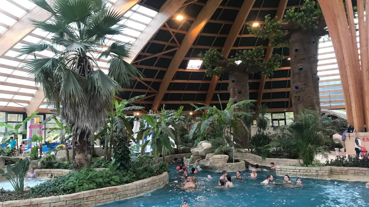 Diversión acuática en Bretaña con niños. Familias disfrutan de una piscina interior tropical con palmeras y toboganes.