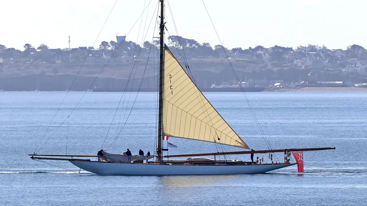 Un élégant voilier, surnommé "mariquita", navigue sur une mer bleue, sa grande voile beige déployée. Une côte verdoyante bordée de maisons se dessine à l'horizon.