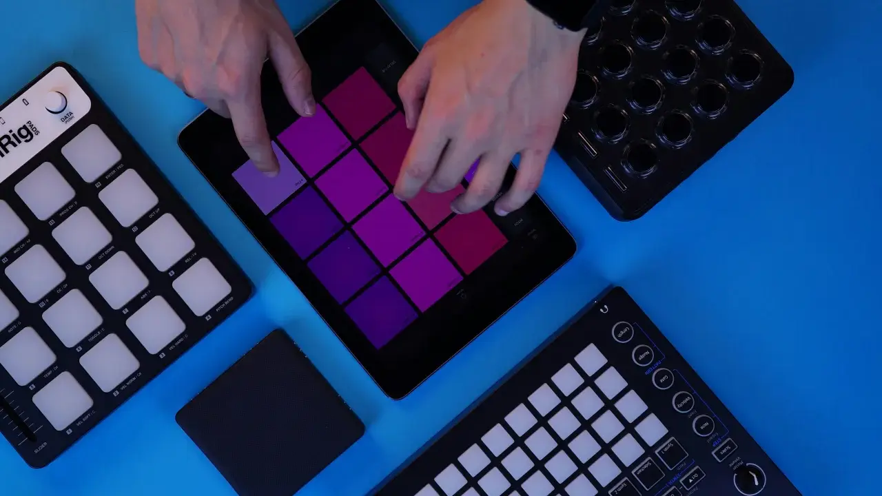 Des mains manipulent une tablette affichant des pads colorés, entourée de contrôleurs midi. Création musicale en cours.