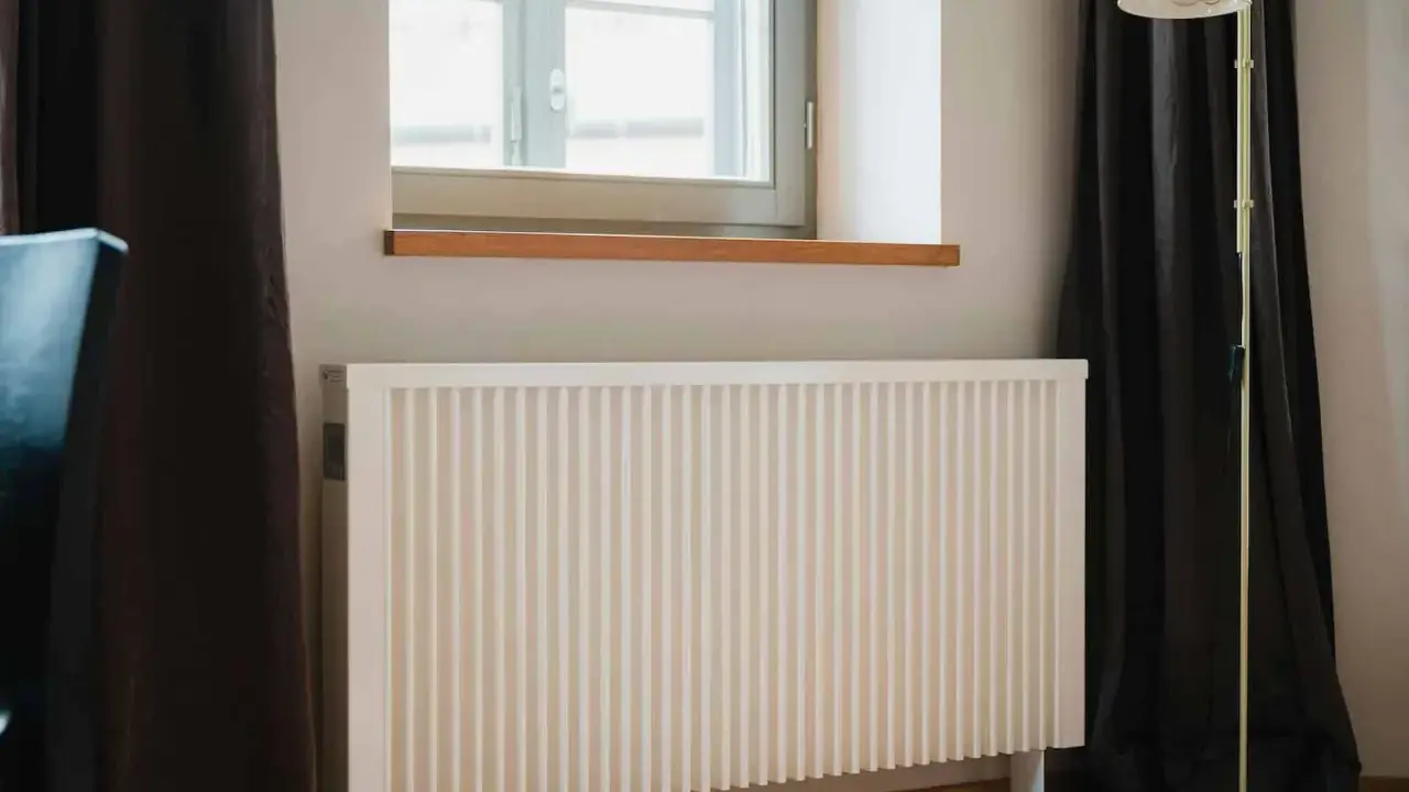 Biały grzejnik pod oknem, obok ciemnej zasłony i lampy stojącej. Dowiedz się, jak ustawić grzejniki przy ogrzewaniu gazowym dla optymalnego komfortu.