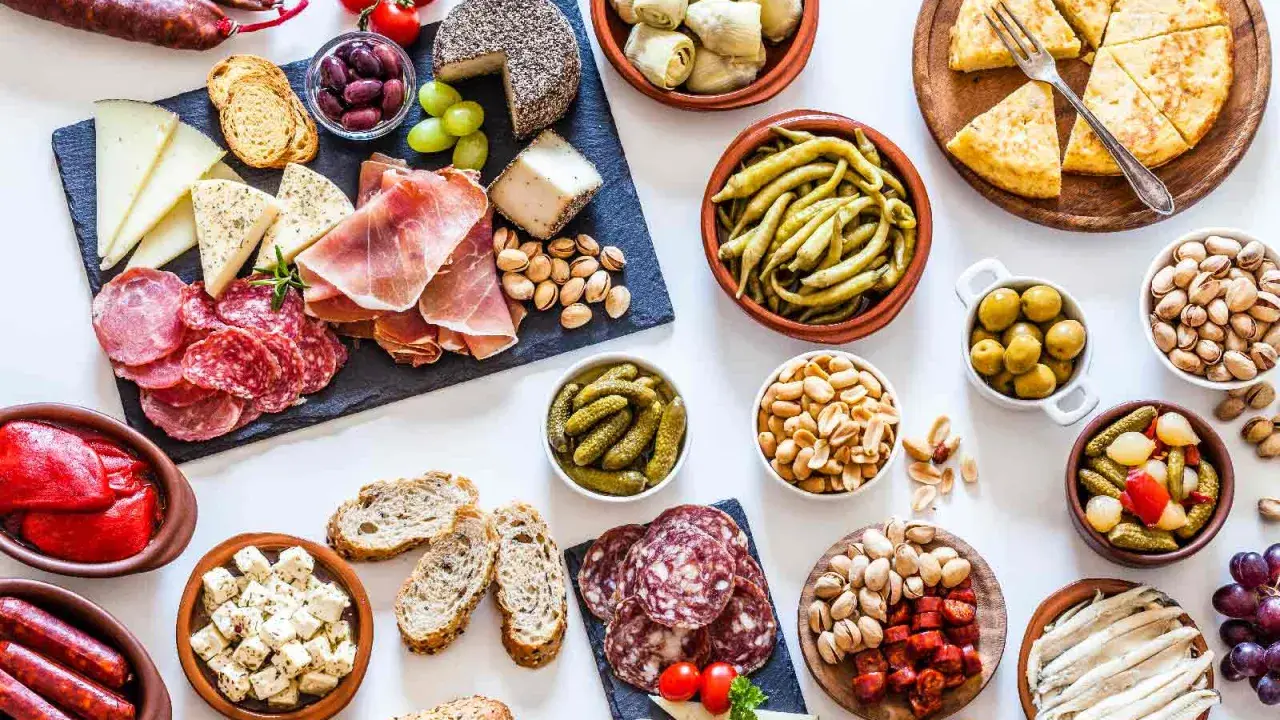 Una mesa llena de aperitivos baratos para mucha gente: embutidos, quesos, aceitunas, frutos secos, pimientos, pepinillos y pan.