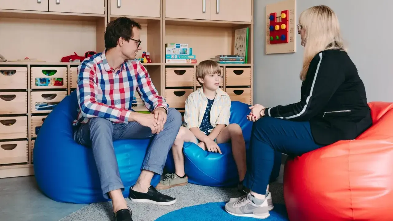 Ojciec, syn i terapeuta dla dziecka rozmawiają w przytulnym pokoju. Na ścianie wisi tablica sensoryczna.