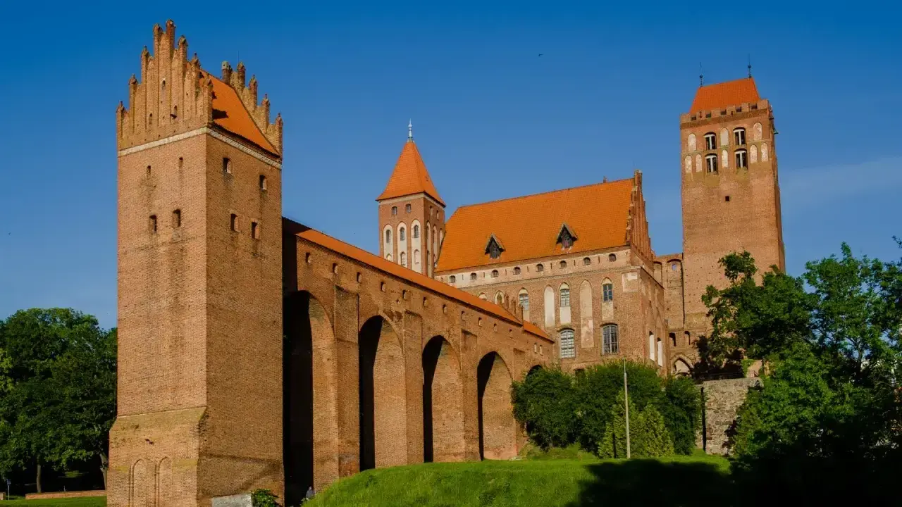 Potężny zamek w Kwidzynie z cegły, z charakterystycznymi wieżami i arkadowym murem obronnym, lśni w słońcu na tle błękitnego nieba.