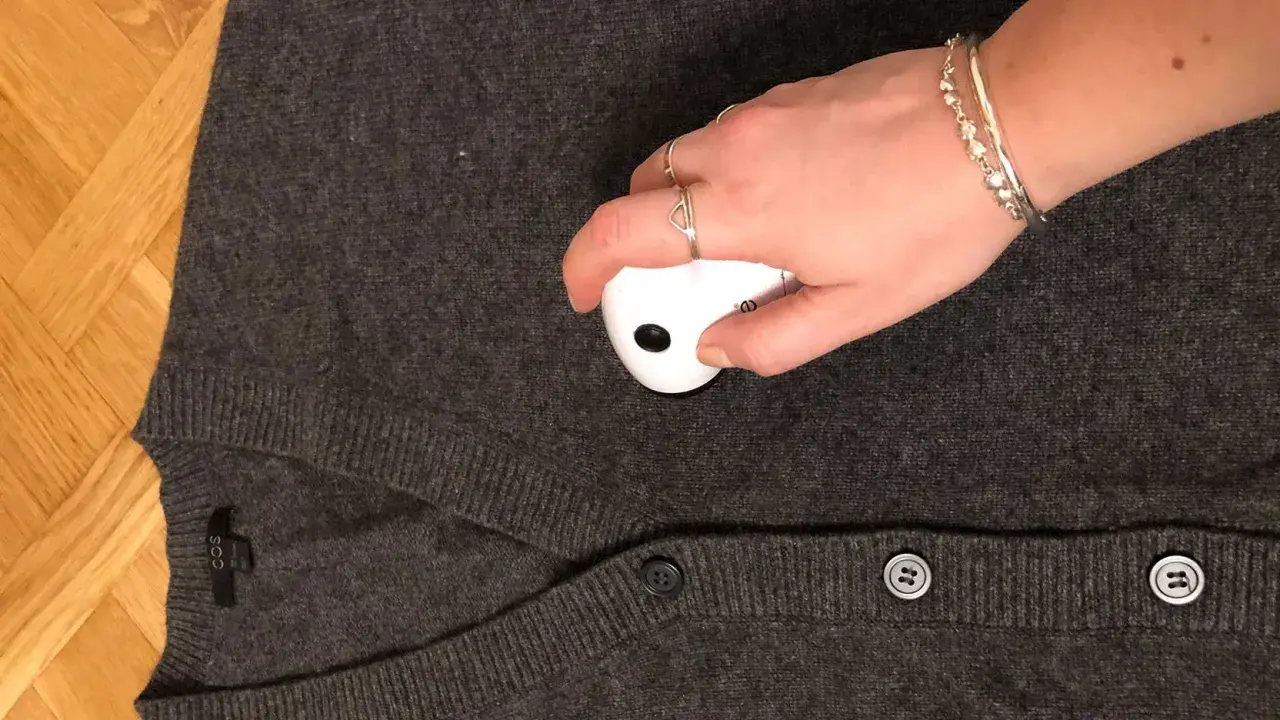 En hand med smycken använder en noppmaskin på en mörkgrå kofta. Materialet känns mjukt, kanske rayon material svenska.
