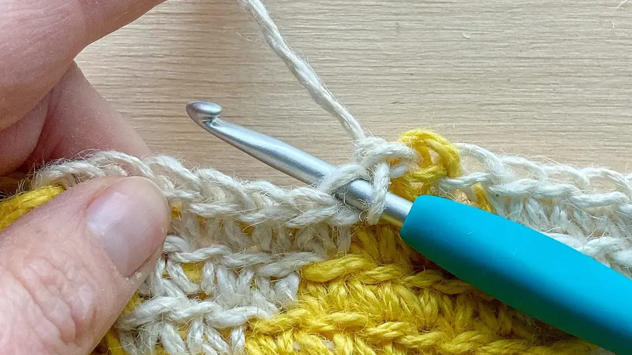 Mano tejiendo con ganchillo, creando un punto bajo con hilo blanco sobre una tela amarilla y blanca.