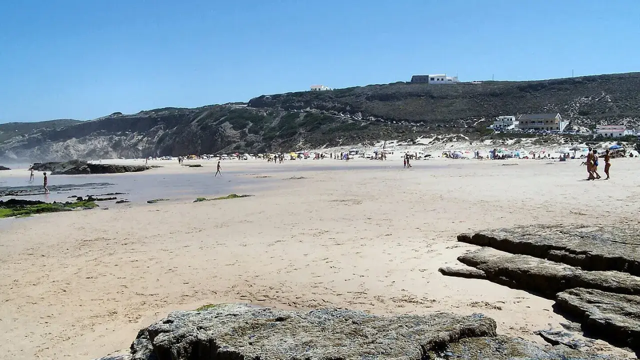 Sonniger Strand bei Monte Clerigo mit Felsen und Menschen, die sich im Wasser und am Ufer aufhalten.